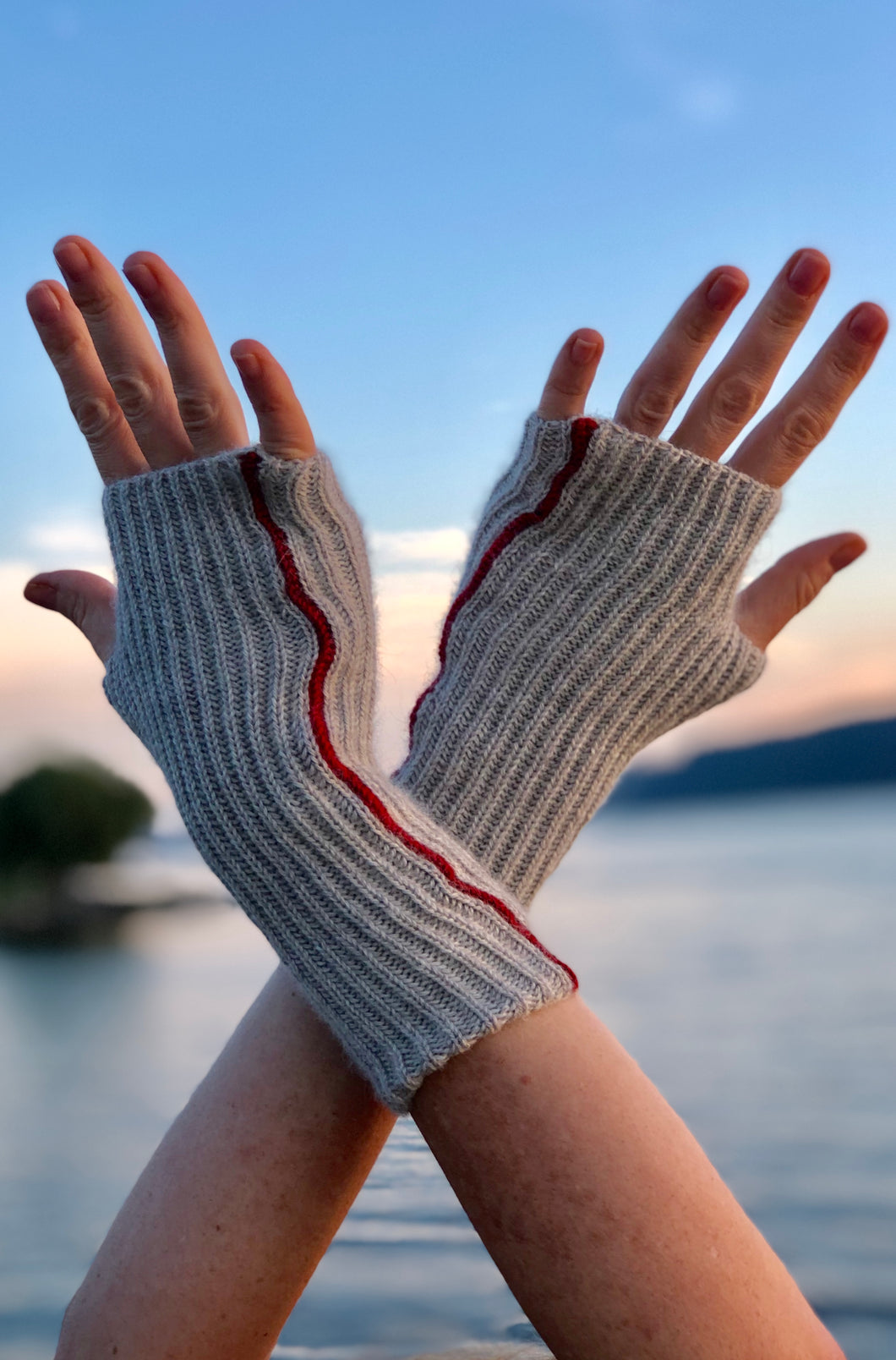 Photo advertising knitting pattern for fingerless mitts. Photo shows two hands extended in the air with a river in the backdrop. The mitts are made with grey alpaca and feature brioche stitches and a red seam.